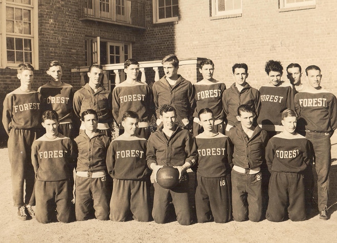 A vintage photo of a boys’ basketball team in “Forest” uniforms posing together outdoors in front of a brick building.