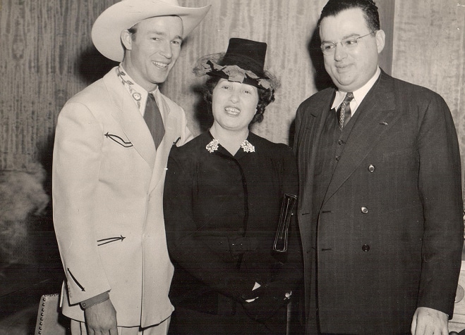 Three people in formal 1940s attire stand together, smiling; one wears a cowboy hat and western suit.