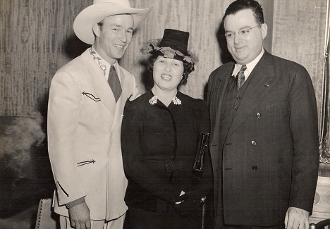 Three people stand together indoors, one in a cowboy hat and suit, one in a dress and hat, and one in a dark suit.