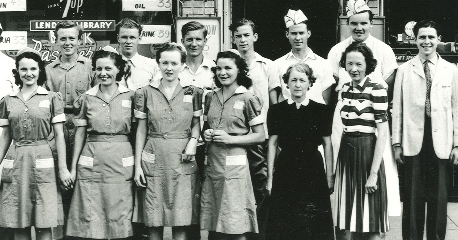 Vintage photo of a group of men and women in uniforms, posing and smiling in front of a storefront.