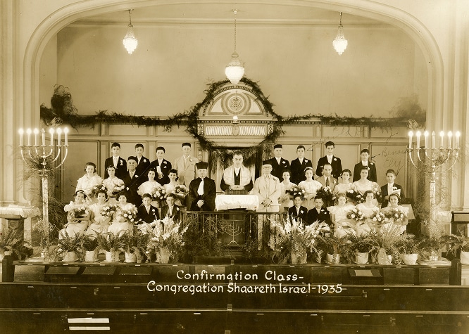 Black-and-white photo of a 1935 confirmation class at a synagogue, with flowers and candlesticks on stage.
