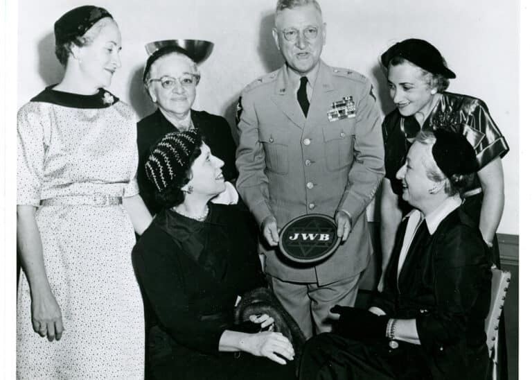 A man in military uniform holds a JWB plaque, surrounded by five women smiling at him in a room.