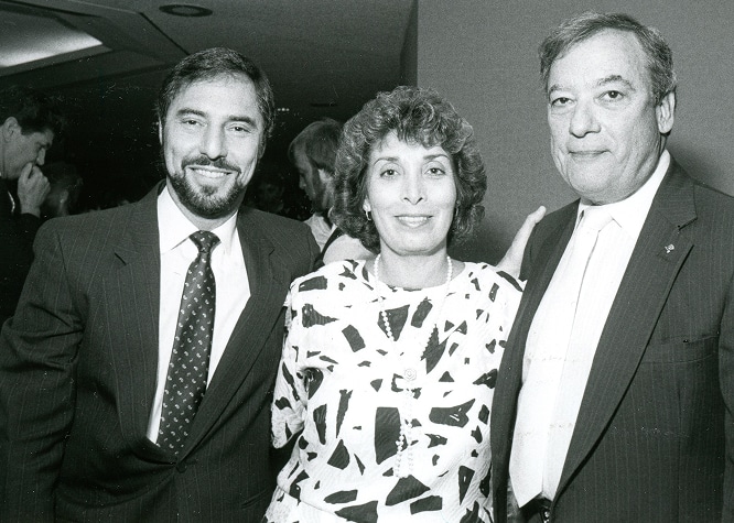 Three adults in formal attire posing together and smiling at an indoor event.