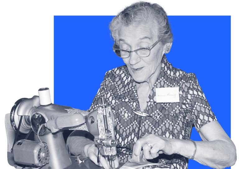 Elderly woman wearing glasses smiles while sewing fabric on a machine, against a blue background.