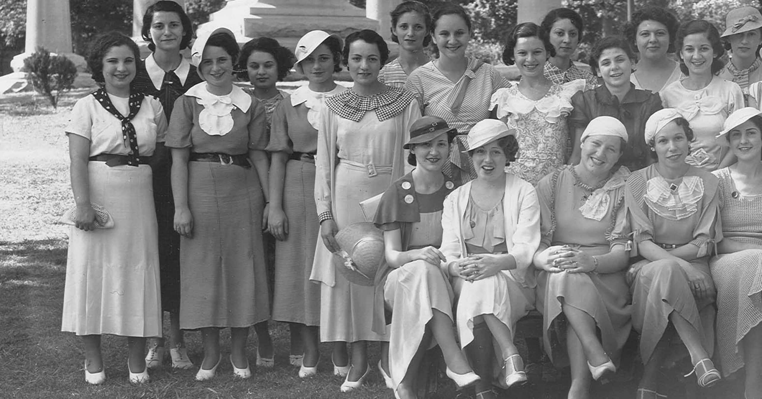 A group of women in 1930s dresses and hats pose together for a photo outdoors, some standing and some sitting.