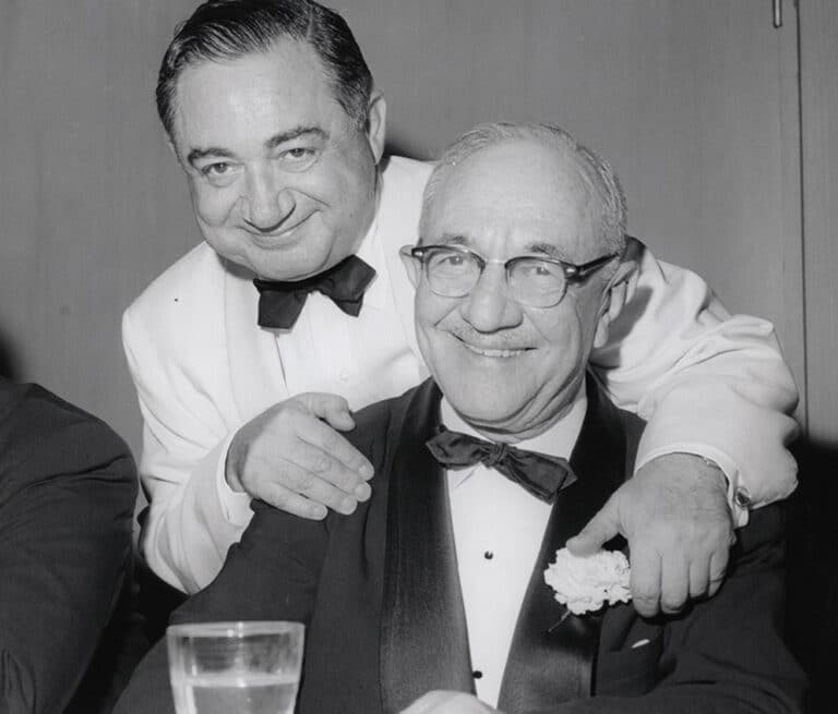 Two older men in tuxedos smile at the camera; one stands behind the other with his hand on his friend’s shoulder.