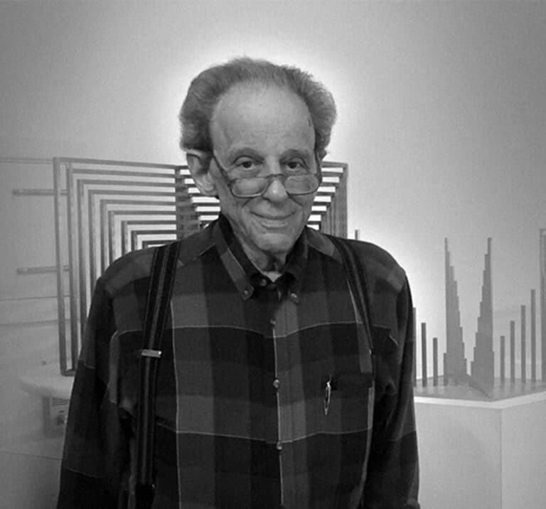 An elderly man in glasses and suspenders stands in an art gallery with geometric sculptures in the background.