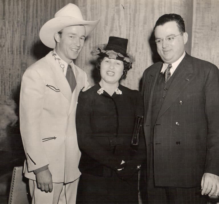 Three people pose together, one man in a cowboy hat, one woman in a hat, and a man in a suit, all smiling.