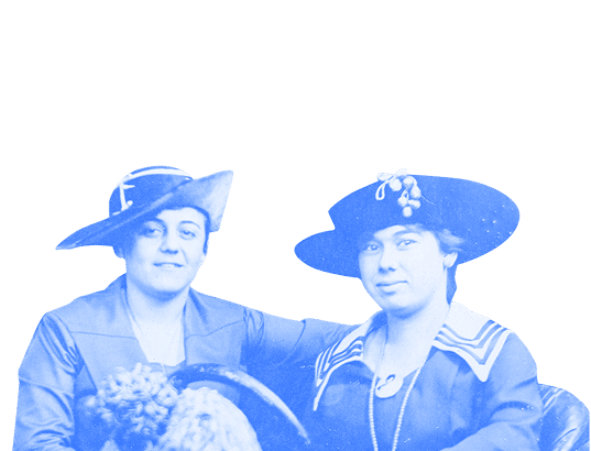 Two women in vintage hats and clothing sit close together, posing for a portrait against a plain background.