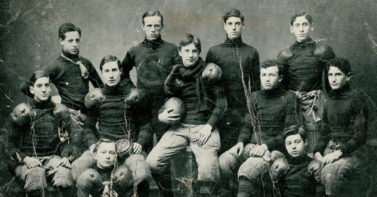 Vintage photo of a football team in old uniforms, posed together holding a football, looking at the camera.
