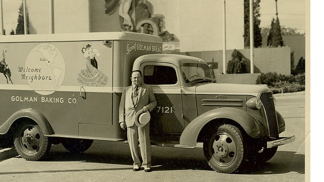 A man in a suit stands beside a vintage Golman Baking Co. delivery truck with “Welcome Neighbors” on its side.