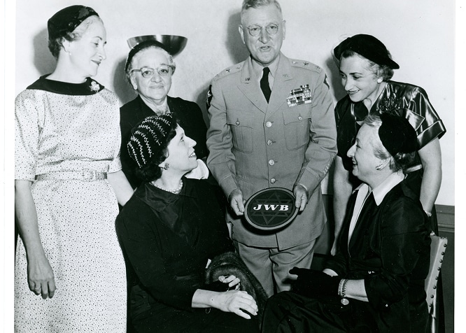 Five women and a man in uniform pose together indoors; the man holds a round object labeled “JWB.”.