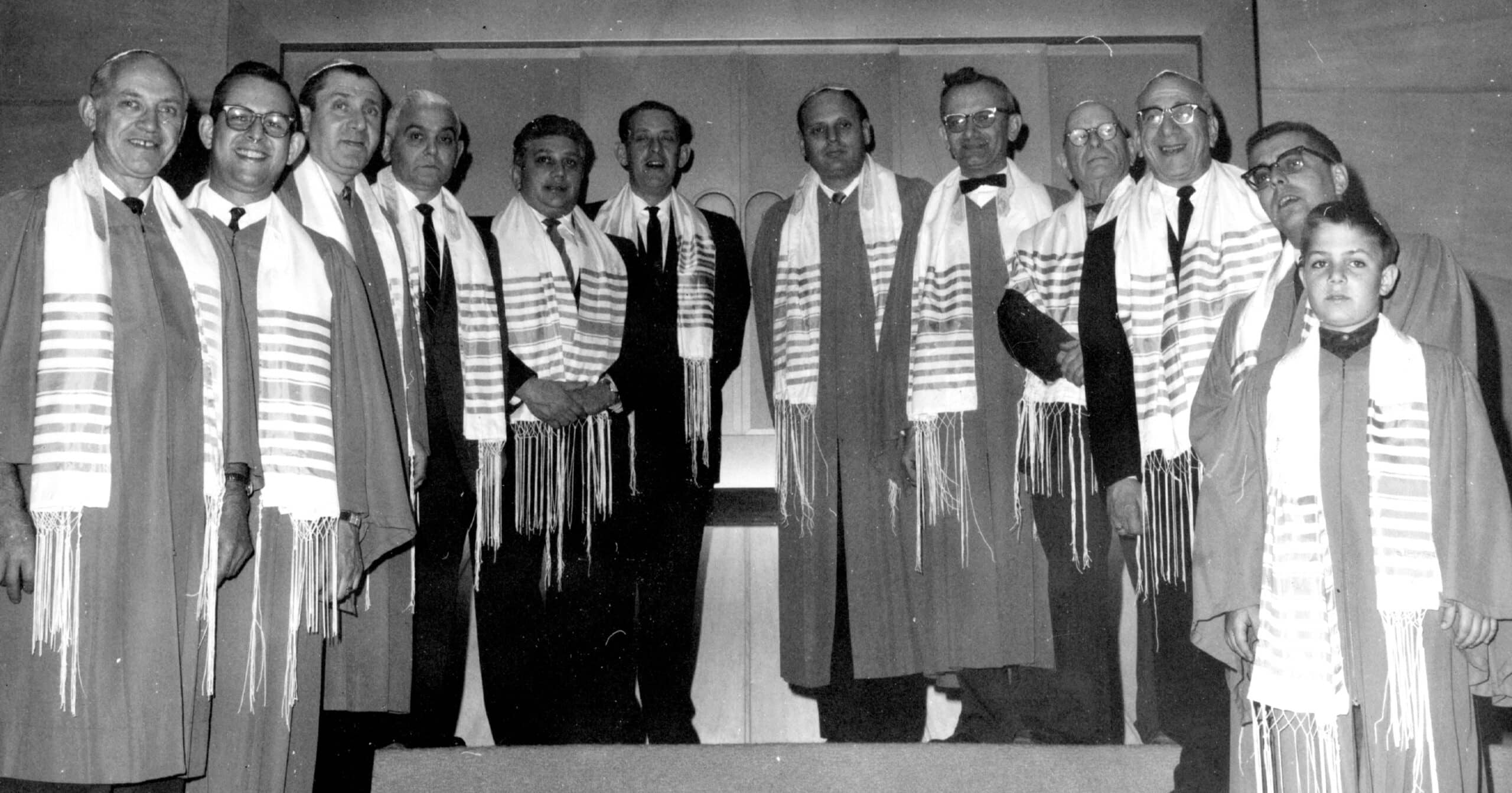 Thirteen people wearing robes and striped scarves stand in a row, facing the camera on a raised platform indoors.