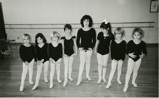 Eight young girls in black leotards and white tights stand in a ballet studio, posing for a group photo.