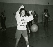 Woman in gym shorts and sweatshirt plays with basketball indoors; several people are in the background.
