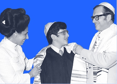 A boy in religious attire smiles as two adults place a prayer shawl on his shoulders against a blue background.