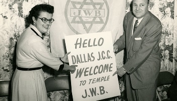 Two people hold a sign reading “Hello Dallas J.C.C. Welcome to Temple J.W.B.” in front of a decorated backdrop.