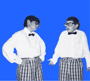 Two people in bow ties and plaid pants, wearing glasses, stand facing each other against a bright blue background.