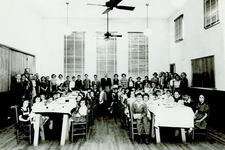 Large group of children and adults gathered around tables in a bright, high-ceilinged room for a formal event.