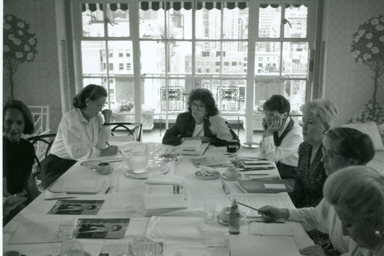 Seven women sit around a table covered with papers, discussing, in a bright room with large windows and city views.