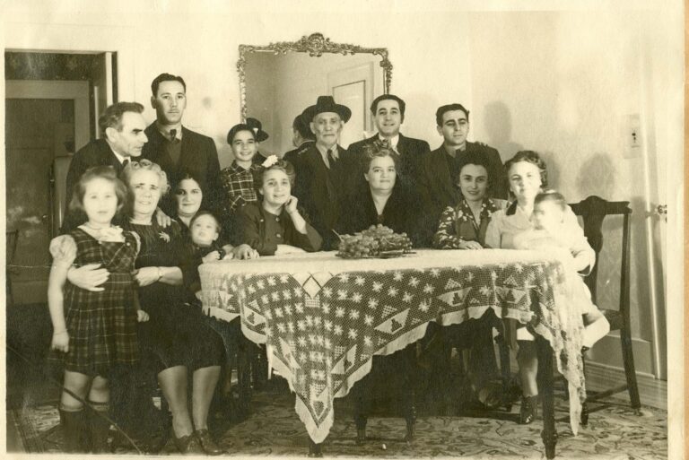 Fourteen people of various ages pose around a table with a patterned cloth in a vintage, sepia-toned room.