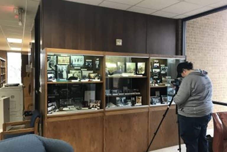 A person uses a camera on a tripod to photograph a glass display case filled with photos and artifacts in a library.