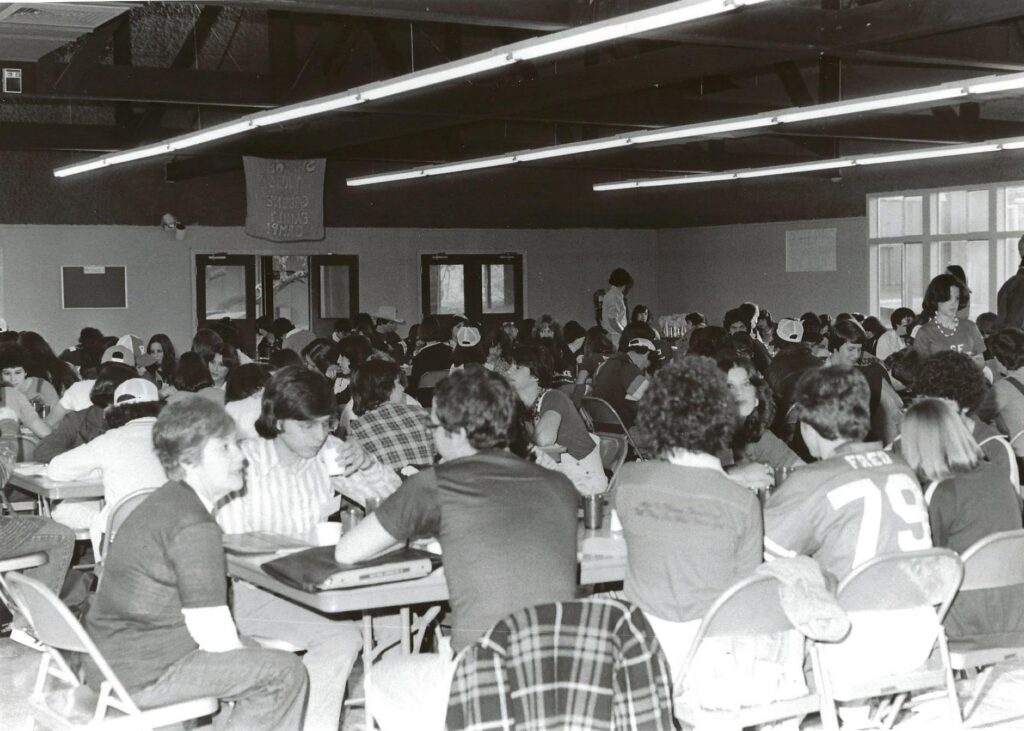 Black and white photo of many people sitting at tables in a crowded cafeteria or dining hall, talking and eating.