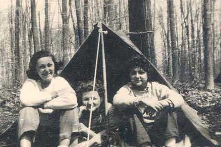 Three people sit in front of a small tent in a forest, smiling, in a black-and-white photo.