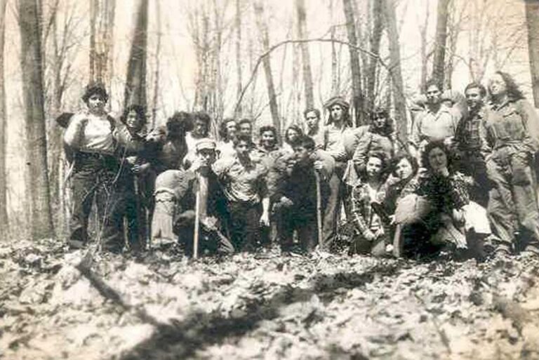 A group of people pose together in a forest, surrounded by bare trees and fallen leaves on the ground.