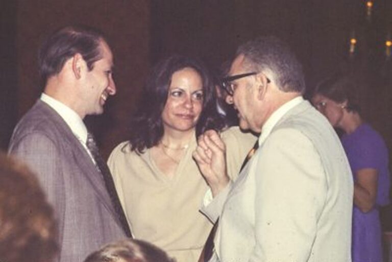 Three people in formal attire are conversing indoors, with another person blurred in the background.