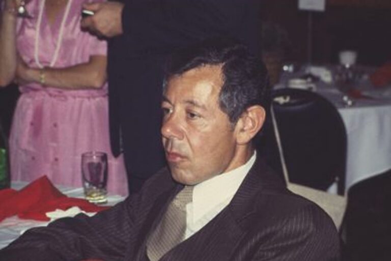 Man in a brown suit sits at a table in a dimly lit room with empty glasses and people in the background.