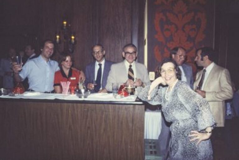 Seven adults in business attire gather around a bar, smiling, with one woman posing playfully in the foreground.