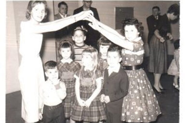 Children stand in a group while two older girls form an arch with their arms at a party. Adults are in the background.