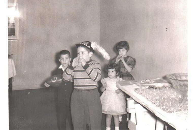 Four children at a party, one in a party hat drinking, others stand nearby, with a table of glasses in front.