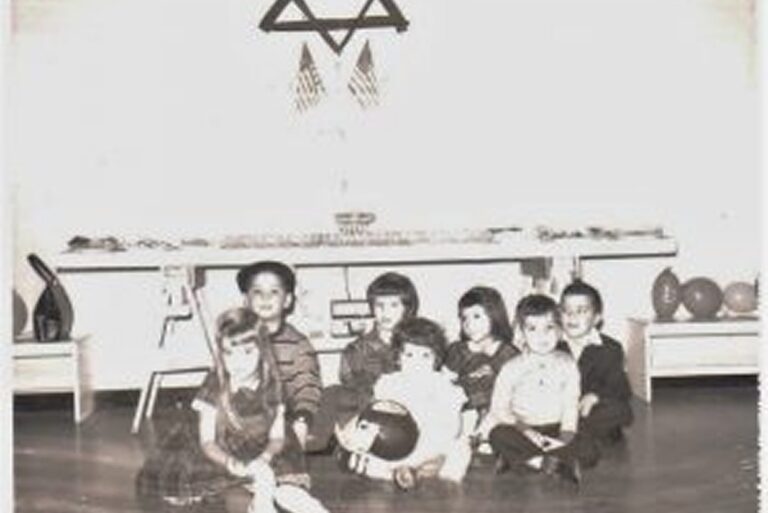 Seven children sit on the floor in front of a table with a Star of David and American flags on the wall behind them.