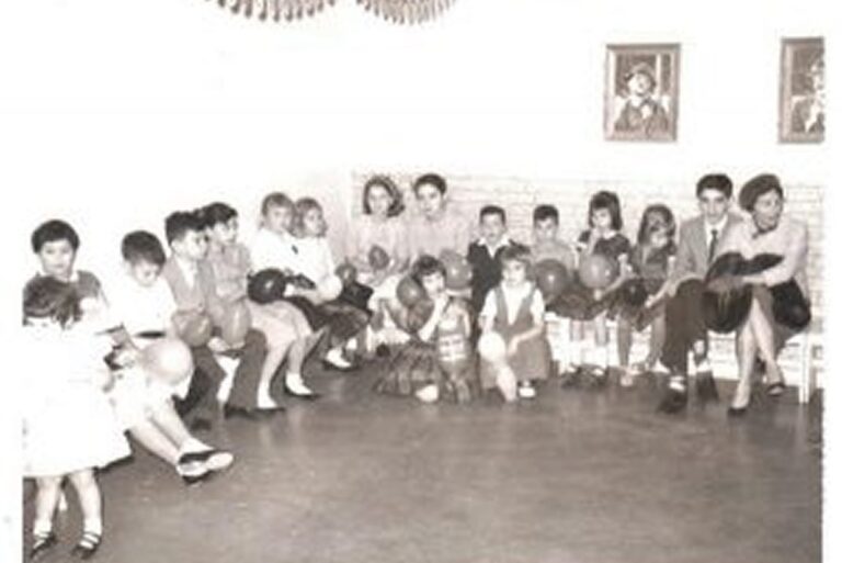 A group of children and adults sit on chairs holding balloons in a room with two framed pictures on the wall.
