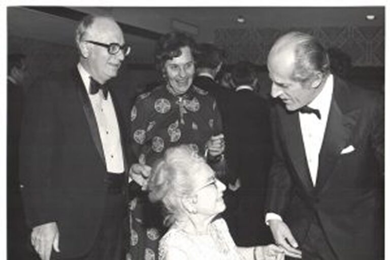 Four adults in formal attire converse at an event; one elderly woman is seated while the others stand around her, smiling.