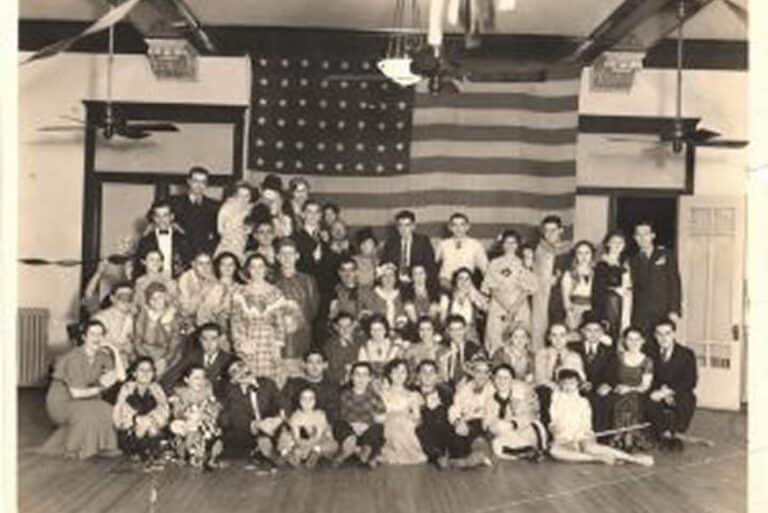 Vintage black-and-white photo of a large group posing indoors in front of a large American flag.