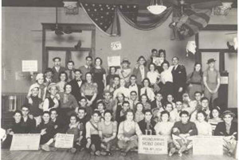 Large group posing indoors, some hold signs; U.S. flags hang behind them; photo appears vintage, likely early 1900s.