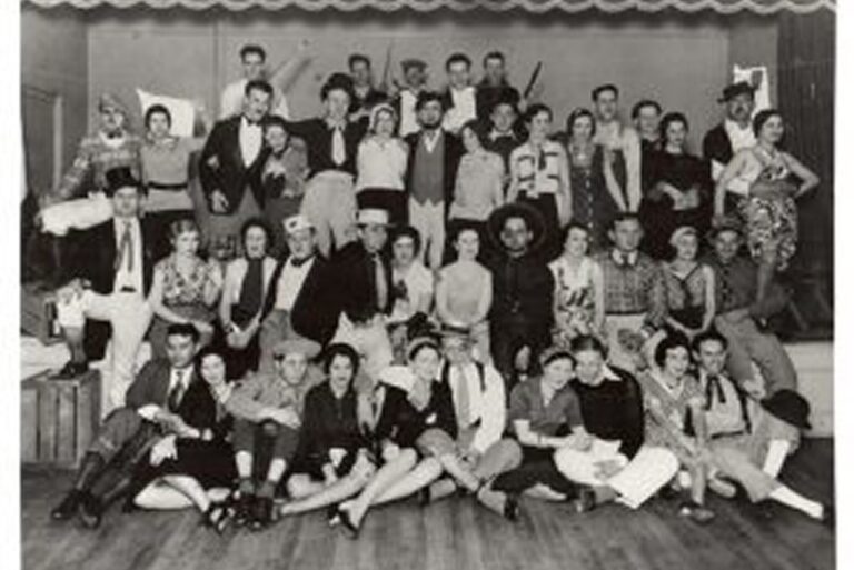 A large group of people in costumes pose on a stage, smiling and sitting or standing in several rows.