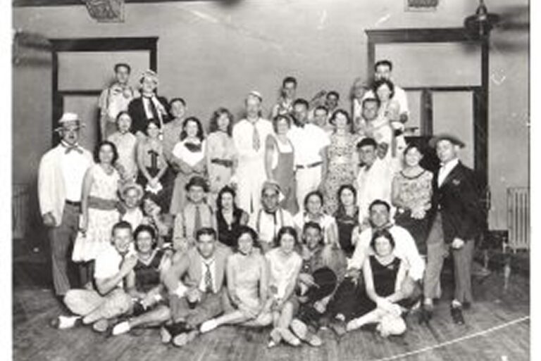 A large group of people in vintage clothing pose together indoors, some standing, others sitting, in a black and white photo.