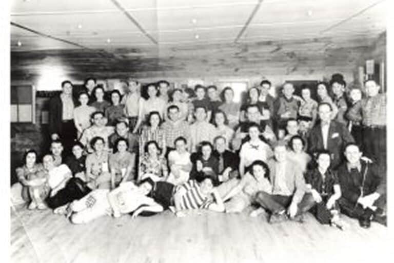Large group of people, men and women, pose together in a hall, smiling, dressed in casual 1940s or 1950s attire.