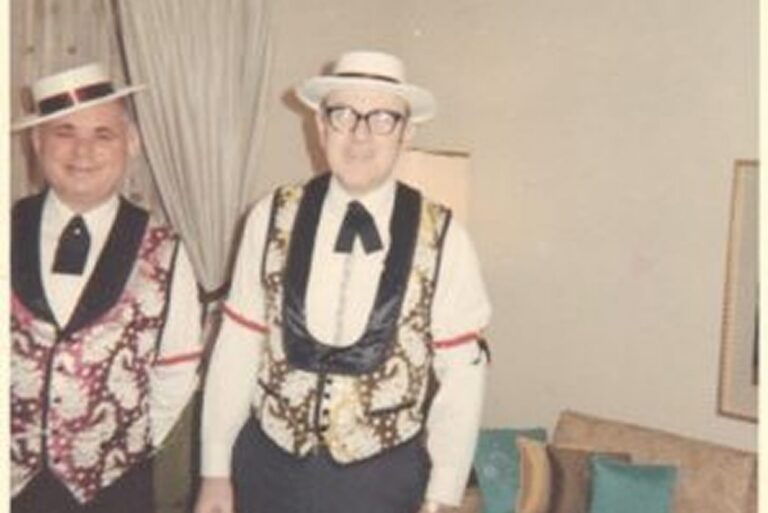 Two men wearing matching vests, bow ties, and hats stand in a living room with a couch and curtains behind them.