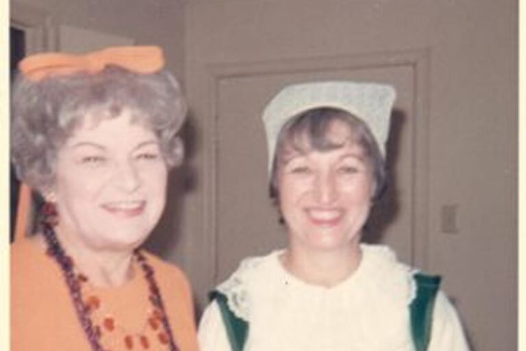 Two women smiling, one in an orange dress with a bow, the other in a white and green outfit with a bonnet.