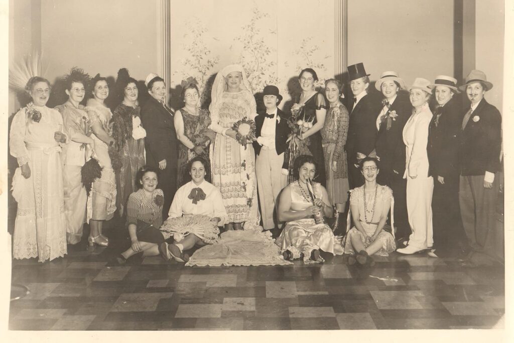 A group of people in vintage costumes pose indoors; some are standing, others are sitting on the floor, all smiling.