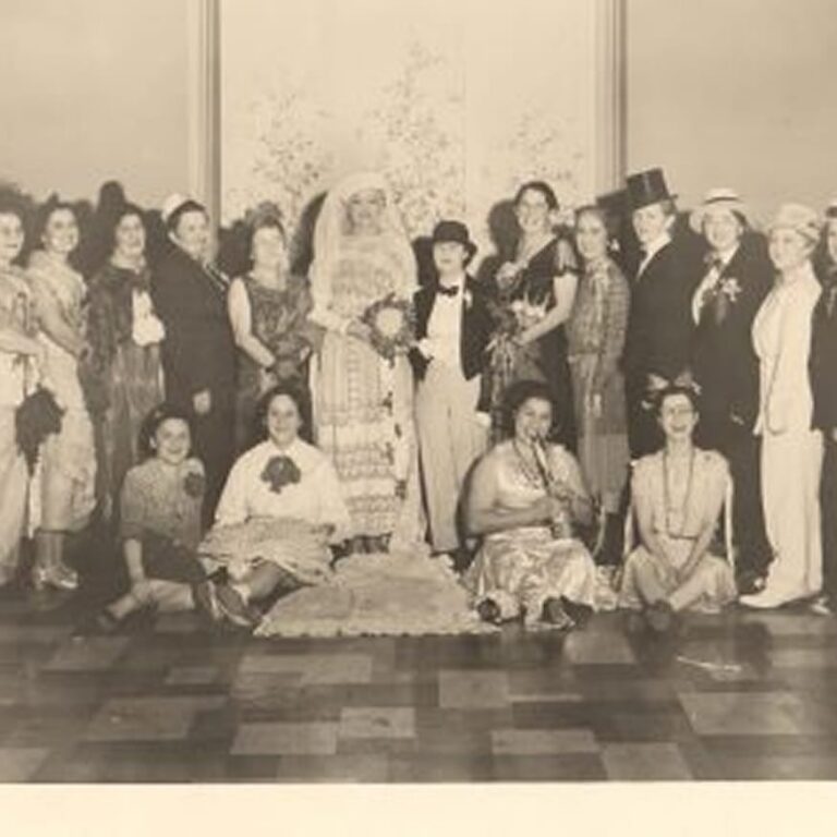 A group of women in costumes pose indoors, some standing, some sitting, with one dressed as a bride in the center.