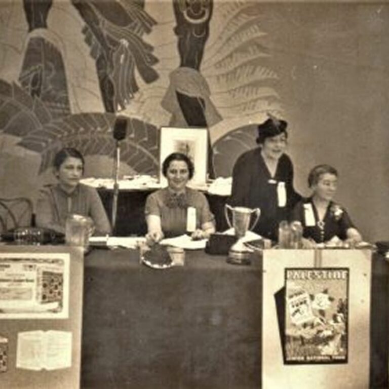 Four women sit at a table with papers, jars, and posters on the front, with a mural in the background.