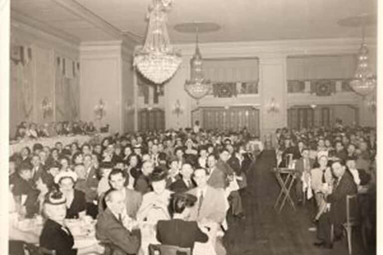 Large crowd seated at tables in an elegant banquet hall with chandeliers, dressed in formal attire.