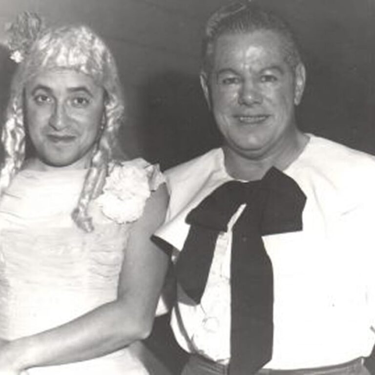 Two men smile; one is in a dress and wig, the other wears a shirt with a large bow. The photo is black and white.