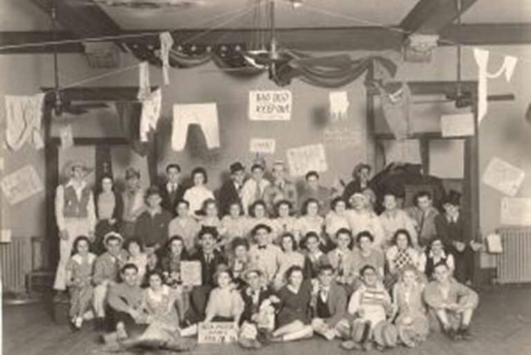Large group of people pose indoors with humorous signs and decorations hanging overhead, in a vintage black-and-white photo.